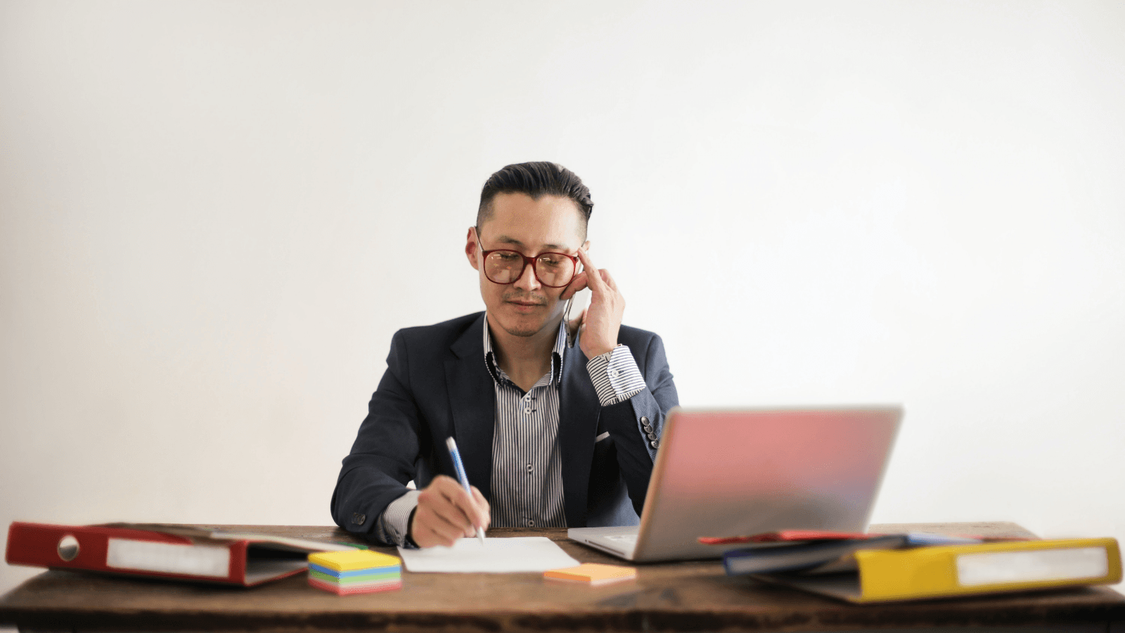 Man in glasses writing at desk next to laptop